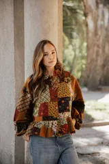 Camisa de estilo patchwork confeccionada en lino, con diseño de bloques de diferentes estampados florales y geométricos en tonos tierra. Presenta cuello camisero, cierre frontal con botones, mangas largas abullonadas con puños elásticos y un ruedo con volado.