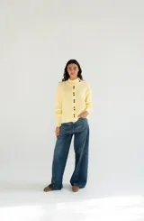 Saco tejido de punto color amarillo claro, con cuello alto asimétrico abotonado, botones oscuros contrastantes en la parte frontal y mangas largas con puños ajustados.