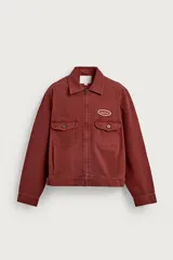Cazadora cropped fit de Zara confeccionada en tejido de algodón con estructura canvas, de color bordó con efecto lavado. Cuenta con cuello solapa, manga larga con puños abotonados, bolsillos frontales de plastrón con solapa, parche bordado en el pecho y cierre frontal con cremallera.