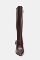 Bota de caña alta en color marrón oscuro, confeccionada en cuero con acabado liso. Presenta un diseño de punta fina, tacón alto fino y una tira decorativa en el tobillo con una hebilla metálica dorada. Incluye cierre lateral mediante cremallera.