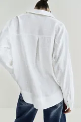 Blusa blanca confeccionada en lino, con cuello polo, escote en V y mangas largas amplias. Presenta un diseño holgado y bajo asimétrico.