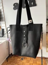 Bolso tipo tote bag negro de cuero con textura granulada, con doble asa de hombro y detalle de tira vertical con tachas y aro metálico.