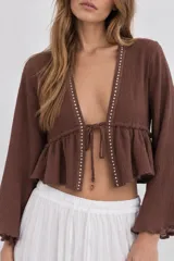 Blusa crop top marrón con escote en V pronunciado, mangas largas acampanadas y cierre de lazo delantero. Tiene detalles de apliques metalizados en el borde del escote y un diseño fruncido en la parte inferior.