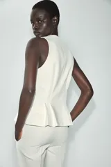 Chaleco entallado blanco con cuello redondo, manga sisa, pinzas estructuradas, detalle de peplum en la parte inferior y cierre frontal con botones dorados.