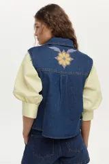 Chaleco de denim azul oscuro con cuello clásico y bordados florales en tonos amarillos, blancos y celestes en la parte delantera.