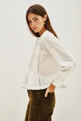 Camisa de algodón blanco con textura de pequeños puntos en relieve, cuello redondo con cierre de botones oculto, detalles de alforzas verticales y encaje en el frente, y mangas largas con puños elásticos.