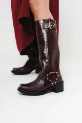 Bota biker de caña alta color bordó con textura símil piel de cocodrilo, hebilla en la parte superior y detalle de aro metálico plateado en la parte inferior. Taco cuadrado y punta cuadrada.