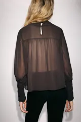 Blusa de cuello subido y manga larga, color marrón oscuro, con detalles de bordados combinados a tono en el canesú y los puños anchos. Cierre en la espalda con abertura y botones.