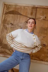 Sweater de punto en color crema con rayas horizontales en tono dorado. Presenta un diseño de cuello redondo con una solapa lateral decorada con botones metálicos dorados y corte holgado.