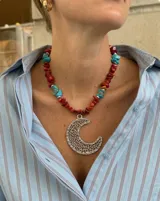 Collar corto de escallas de piedra coral en tonos rojizos, intercaladas con piedras turquesas y pequeños apliques plateados. Cuenta con un gran dije colgante de metal plateado con diseño de media luna texturada y calada.