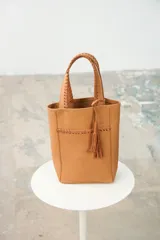 Matera tipo bolso de mano color tostado, confeccionada en cuero con detalles de costuras bordadas a mano y flecos colgantes decorativos.