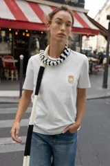 Camiseta negra de algodón, corte regular, con cuello redondo ribeteado en blanco y un bolsillo frontal con un broche metálico decorativo dorado.