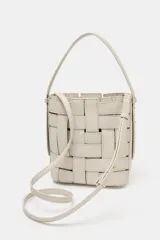 Bolso tipo bucket de diseño trenzado en color marrón oscuro con costuras a la vista en contraste. Cuenta con asa de mano fija, correa bandolera extraíble y compartimento interior de tela en color crudo con cierre mediante fruncido.