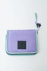 Billetera plegable color lila con ribetes y cierre en color celeste. Posee un parche cuadrado negro con texto blanco en el frente. Incluye cordón ajustable con tope negro y terminales celestes.