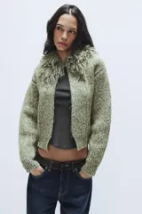 Chaqueta de punto de textura jaspeada en color verde, con cuello redondo adornado con flecos del mismo tejido, manga larga y cierre frontal con corchete metálico.