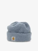 Gorro tipo beanie de la marca Carhartt, confeccionado en tejido de pelo sintético tipo sherpa de color azul grisáceo. Presenta un diseño de puño doblado con una etiqueta rectangular tejida que incluye el logo de la marca.