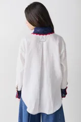 Camisa de lino blanco con cuello y puños tejidos a mano en crochet de color azul con borde rojo. Presenta cierre frontal con botones, bolsillo lateral y costuras a la vista en contraste.