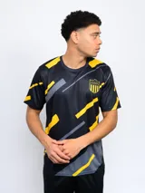 Remera de entrenamiento Fynar del Club Atlético Peñarol, color negro con estampado geométrico en amarillo y gris. Presenta cuello redondo, mangas cortas y el escudo del club en el pecho.