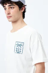 Remera blanca de corte amplio con mangas cortas y estampado de una postal con una palmera y el mar.