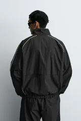 Chaqueta técnica de corte relajado en color verde oscuro, con cuello subido, cierre frontal de cremallera y detalles de líneas en contraste. Presenta bolsillos frontales con cremallera oculta y acabados elásticos en puños y ruedo.