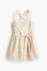 Vestido corto beige con detalles de broderie en el escote y en el ruedo. Cuenta con escote cuadrado, breteles anchos cruzados en la espalda, cintura fruncida con lazo para atar y falda con vuelo.
