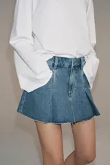 Falda corta de tiro medio confeccionada en tejido denim de algodón. Presenta diseño con tablas, bolsillos delanteros, cintura con trabillas y bajo desflecado. Cierre frontal con cremallera, botón y ganchos metálicos.