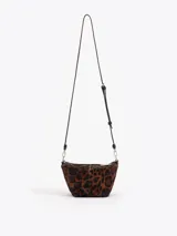 Bandolera de tamaño pequeño confeccionada en pelo vacuno con estampado animal print de leopardo y detalles en cuero vacuno flor. Presenta una cadena metálica decorativa en el frente, aplique metálico con logo JCH y correa de cuero ajustable.