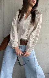 Camisa de manga larga color beige con detalles de encaje y calados verticales en el frente y las mangas, cuello en V y cierre frontal con botones.