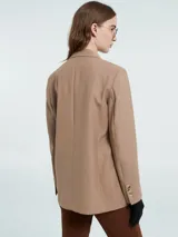 Blazer sastrero color tostado con solapas clásicas, cierre frontal de doble botonadura y bolsillos con solapa. Presenta mangas largas con botones decorativos en los puños.