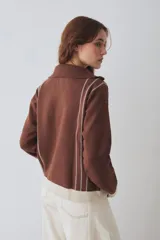 Buzo de tejido de punto color marrón con cuello alto y cierre frontal parcial. Presenta rayas verticales finas en color claro, puños y ruedo en tejido acanalado color crema, y detalles en color verde lima en el tirador del cierre, el cordón de ajuste del ruedo y el parche bordado en el pecho.