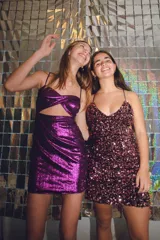 Vestido corto de fiesta color plateado con acabado brillante. Presenta un diseño de dos piezas que simula un conjunto de top y pollera, con aberturas laterales en la cintura, escote en V y tirantes finos que se cruzan en la espalda.