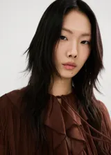 Blusa de color marrón chocolate, confeccionada en tejido plisado con textura. Presenta un diseño de cuello redondo con detalle de lazo, mangas largas amplias con puños acampanados y un corte holgado con volados sutiles en la parte frontal.