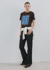 Camiseta marrón de algodón con cuello redondo y manga corta, con estampa frontal azul de flores y texto "Bloom More Worry Less".