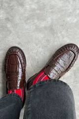 Mocasín de cuero marrón con textura croco y acabado brillante, de diseño clásico con detalle de antifaz en el empeine y costuras pespunteadas alrededor de la puntera.