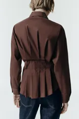 Camisa de manga larga en color celeste claro, confeccionada en tejido de popelín. Presenta cuello solapa, cintura entallada con detalle de pliegues y cierre frontal con botones ocultos por tapeta.