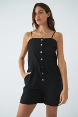 Monoprenda corta de color verde oliva, con tirantes finos ajustables, cierre frontal con botones y cintura elástica con cordón ajustable. Cuenta con bolsillos laterales.