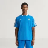 Camiseta de fútbol Adidas del Real Madrid, inspirada en los años 80. Diseño de color azul con finas rayas verticales blancas, cuello en V blanco, mangas cortas con las tres tiras características de la marca y escudo retro del club bordado en el pecho.