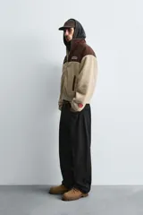 Cazadora de corte relajado confeccionada en tejido de borreguillo color beige combinado con tejido técnico marrón en la parte superior. Presenta cuello subido, cierre frontal con cremallera, bolsillos laterales de vivo y puños y bajo elásticos. Incluye parches y bordados decorativos en el pecho.