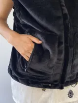 Chaleco de peluche negro con cuello alto, cierre frontal, bolsillos delanteros y detalles decorativos en los hombros.