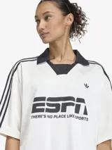 Camiseta de fútbol de corte oversize, color blanco con detalles en negro. Presenta cuello tipo polo con escote en V, mangas cortas con las icónicas tres tiras de Adidas en los hombros y estampado gráfico frontal inspirado en la estética deportiva de los años 90.