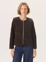 Chaqueta de paño color marrón oscuro, de corte clásico y cuello redondo, con cierre frontal dorado. Presenta bolsillos rectangulares con ribetes en color rojizo y parches de pana a rayas en los hombros.