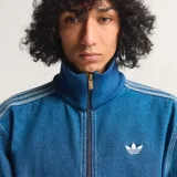 Campera deportiva Adidas Originals modelo Firebird, confeccionada en denim azul. Presenta cuello alto, cierre frontal, puños y cintura de tejido acanalado azul oscuro, y las tres franjas características de Adidas en las mangas en tono más claro. Incluye el logo del trébol bordado en blanco en el pecho.