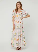 Vestido largo confeccionado en una mezcla de viscosa, algodón y lino con estampado de frutas y soles sobre un fondo rosado pálido. Presenta cuello en V, mangas de largo medio con puños elásticos y un cierre frontal completo con botones.