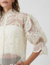 Blusa de tul bordado color beige, marca Liv Los Angeles, con cuello alto, cierre frontal de botones y mangas ¾ amplias con volados.