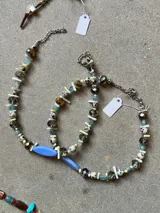 Collar artesanal compuesto por una combinación de cuentas de cristal facetado en tonos gris y bronce, piedras tipo dalmata, cuentas cilíndricas de color crema y cuentas de vidrio azul claro. Presenta un dije central alargado de color azul pastel y terminaciones en cadena de acero.
