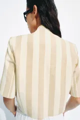 Blazer cropped de manga corta de Zara Woman Collection, confeccionado en hilatura con algodón, con diseño a rayas verticales en tonos beige y blanco. Cuenta con cuello subido, bolsillos de vivo en el delantero, forro interior y cierre frontal con botón.