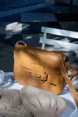 Cartera tipo bandolera de cuero con textura grabada, color beige. Presenta un detalle de herraje metálico dorado en la solapa frontal y placa con logo de la marca. Incluye correa ajustable para llevar al hombro o cruzada.