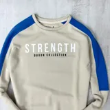 Buzo color arena con mangas ranglan azul eléctrico y estampa frontal con la palabra "Strength" y la frase "Duxon Collection".