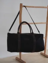 Bolso de viaje tipo duffel de lona negra con detalles en cuero tostado, asas de mano y correa ajustable para el hombro.