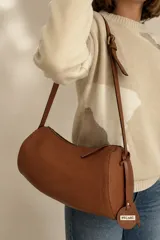 Cartera cilíndrica de cuero color suela, con cierre superior y correa larga ajustable.